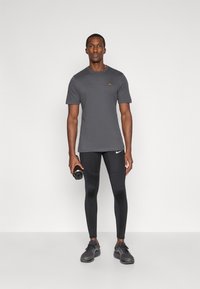 Pilkas trumpas rankovės marškinėliai su mažu logotipu, derinami su juodomis aptemptomis pėdkelnėmis ir juodais sportiniais bateliais, laikomais juoda vandens buteliu.