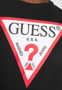 Camiseta de algodón negra con un gran logo triangular en blanco y rojo que incluye "GUESS U.S.A", un signo de interrogación central y el texto "WASHED JEANS".
