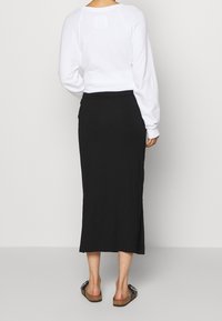 Une personne portant une chemise blanche à manches longues, une jupe noire mi-mollet avec une fente sur le côté et des sandales noires à enfiler, se tenant contre un fond uni.