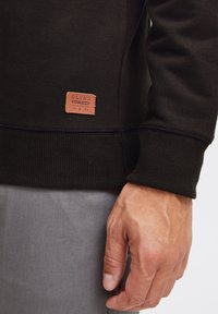 Sweat-shirt noir en matériau doux avec des poignets côtelés, muni d'une étiquette en cuir beige marquée "BLEND" en bas à gauche. Un pantalon gris est partiellement visible.