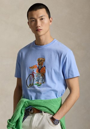Joven con camiseta azul claro con un gráfico de oso, sosteniendo una bicicleta roja, pantalones claros, suéter verde atado a la cintura y reloj de pulsera.
