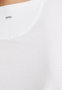 Vit bomull t-shirt med rund halsringning och en liten "RAW" logotyp på baksidan, slät textur med lätt variation i tyget.