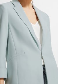 Ljusblå blazer i slätt tyg. Utrustad med spetsslag, enkel knäppning och sidofickor med uppvikt ärm.