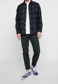 Zelená a tmavě modrá károvaná košile z bavlněné směsi, zapínání na knoflíky. Kombinováno s tmavě zelenými kalhotami a fialovými teniskami Vans s bílými podrážkami.