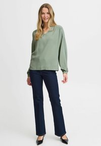 Blusa verde chiaro con scollatura a V, maniche lunghe e tessuto texturizzato, abbinata a jeans a campana blu scuro e décolleté neri a punta.