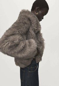 Kunstpelzjacke in Grau mit hohem Kragen, voluminösen Ärmeln und kurzem Schnitt. Kombiniert mit dunklem Denim, der Struktur und Fülle betont.