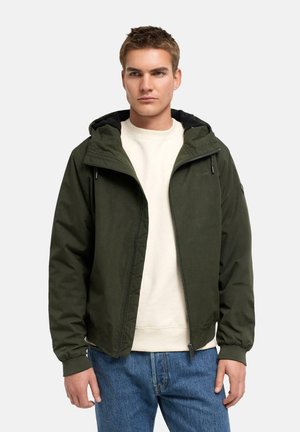 Jeune homme portant une veste à capuche vert foncé par-dessus un sweat-shirt blanc et un jean bleu, debout devant un fond clair uni.