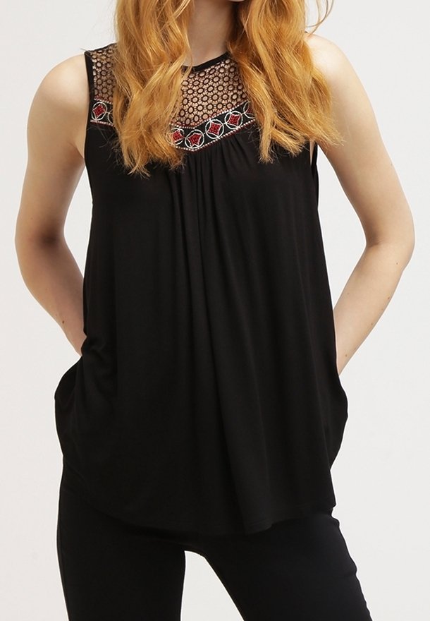 Femme portant un top noir sans manches avec dentelle florale et détail brodé rouge au niveau de l'encolure, mains dans les poches et pantalon noir.