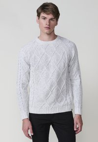Witte, gebreide trui met een ruitpatroon, ronde halslijn en geribbelde manchetten en zoom. Draagt het met zwarte broek, wat de gestructureerde stof laat zien.