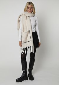 Écharpe en laine crème avec des franges, présentant une poche attachée bordée de noir. Portée sur un haut blanc ajusté et une jupe noire, accompagnée de bottes noires.