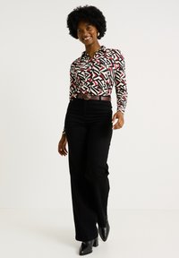 Pantalons larges noirs associés à une chemise à manches longues avec un motif géométrique en noir, blanc et rouge. Ceinture marron et escarpins noirs.