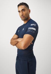 Tamsiai mėlynas sportinis marškinėlis su prigludusia juosta ir trumpomis rankovėmis, ant kairiojo rankovės esantys UFC ir Venum logotipai balta spalva.