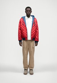 Κόκκινο καπιτονέ μπουφάν με μπλε κουκούλα, άσπρη μπλούζα, μπεζ cargo παντελόνι και γκρι παπούτσια με υφασμένη σόλα. Το μπουφάν διαθέτει ριγέ μανίκια.