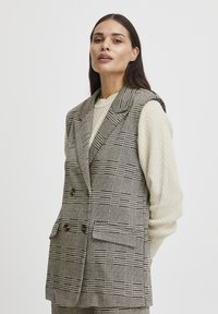 Blazer sans manches à carreaux en gris et blanc, doté de boutons double boutonnage et de poches avant, superposé sur un pull texturé crème.