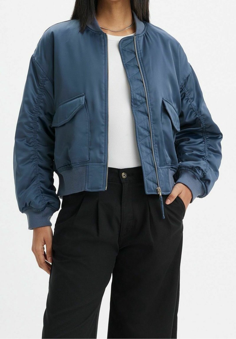 Personne portant une veste bomber bleue avec des poches avant sur un haut blanc et un pantalon noir, main dans la poche, sur un fond uni.