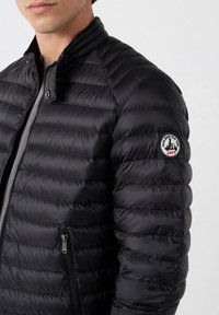 Giubbotto invernale nero con design trapuntato, colletto alto, zip frontale e tasche laterali. Presenta un badge tondo sul maniche sinistra.
