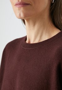 Femme portant un pull à col rond marron foncé et une boucle d'oreille argentée en forme d'anneau, visible du cou à la moitié de la poitrine sur un fond clair.