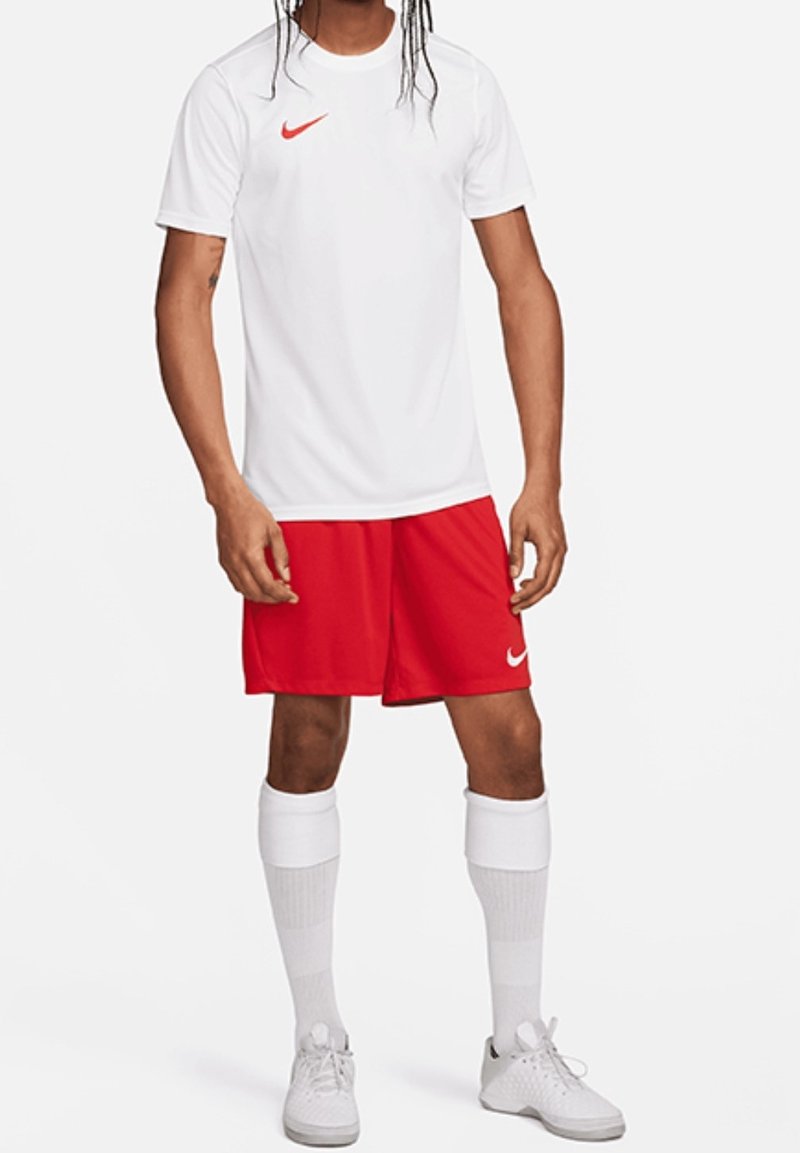 Personne portant un t-shirt de sport blanc avec un signe rouge, un short de sport rouge avec un signe blanc, des chaussettes blanches montantes et des baskets blanches, debout.