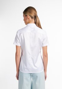 Chemise à manches courtes blanche, en tissu lisse. Dispose d'un col montant, de poignets retournés et d'une couture centrale dans le dos. Coupe ajustée.