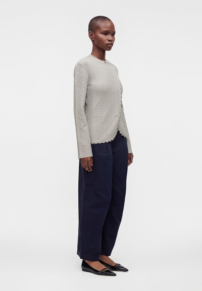 Personne debout de profil portant un haut beige à manches longues, un pantalon large bleu marine et des ballerines noires sur un fond blanc uni.