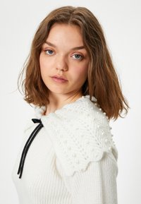 Weißer Strickpullover mit einem gewellten Kragen, der mit Perlen und einem schwarzen Schleifenband verziert ist. Das strukturierte Gewebe vermittelt einen gemütlichen Eindruck.