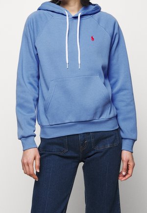 Personne portant un sweat à capuche bleu clair avec des cordons blancs et un logo rouge, associé à un jean bleu foncé, debout devant un fond uni.