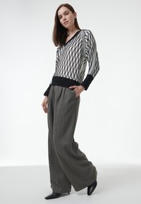 Schwarzer und weißer, diamantförmiger, gestrickter Pullover mit V-Ausschnitt, kombiniert mit weit geschnittenen, grauen, strukturierten Hosen und schwarzen, spitzen Absätzen.