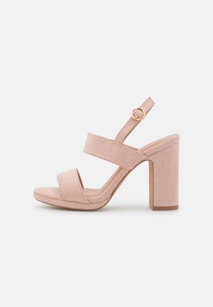 Beige ruskindssandal med høje hæle, to remme, en justerbar anklerem med guldfarvet spænde og en kraftig blokhæl.
