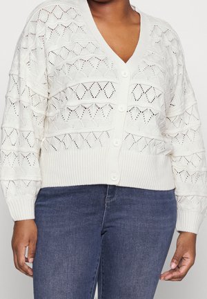 Frau trägt eine weiße Strickcardigan mit Knöpfen und Rautenmuster sowie eine blaue Jeans, abgebildet von der Brustmitte bis zur Hüfte.