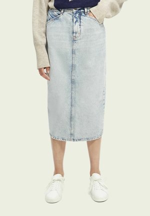 Persoon die een lichtblauwe denim midi-rok met voornaad, een beige trui en witte sneakers draagt, staand tegen een effen achtergrond.