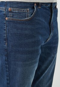 Jeans de mezclilla azul oscuro con corte recto, cinco bolsillos y remaches de cobre. Textura suave con desvanecimientos sutiles a lo largo de las costuras.