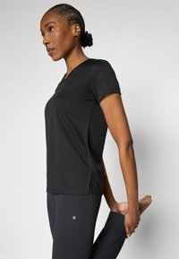 Melnā, īsroku sporta t-krekls ar V-izgriezuma dizainu, ar smalku logotipu sānos. Audums izskatās gluds un viegls.