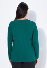 Dlouhý rukáv top v tmavě zelené látce, s kulatým výstřihem. Hladká textura s mírným leskem, volný střih a oblý lem.