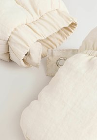 Beige pufferjacka i material med quiltad textur, som har en tryckknappsstängning med en silverögl på ena sidan och samlade tygkanter.