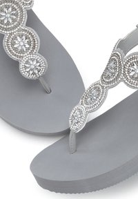 Grijze flip-flops met een gestructureerde zool, een decoratieve band met ronde versieringen en strassaccenten in een bloemontwerp.