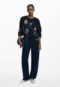 Black knitted sweater with blue and yellow floral embroidery, paired with wide-leg glittery dark blue jeans and white slip-on shoes.