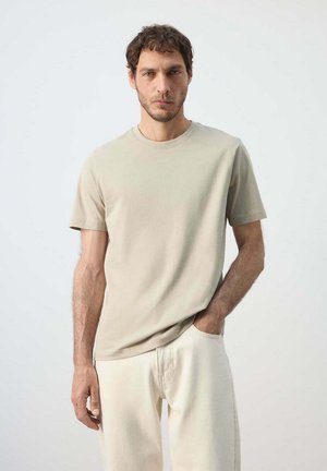 Mann mit kurzen lockigen Haaren, der ein beiges kurzärmliges T-Shirt und helle Hosen trägt, steht mit einer Hand in der Tasche.