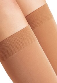 Cosce che indossano collant compressivi beige con bande elastiche testurizzate nella parte superiore su uno sfondo di pelle chiara.
