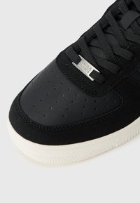 Black sneaker toe with perforated leather, suede trim, black laces, white sole, and silver lace tag engraved with brand name.