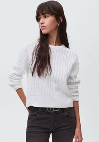 Weißer Strickpullover mit strukturiertem Muster, Rundhalsausschnitt und lockerer Passform. Kombiniert mit dunklen Jeans und einem schwarzen Gürtel.