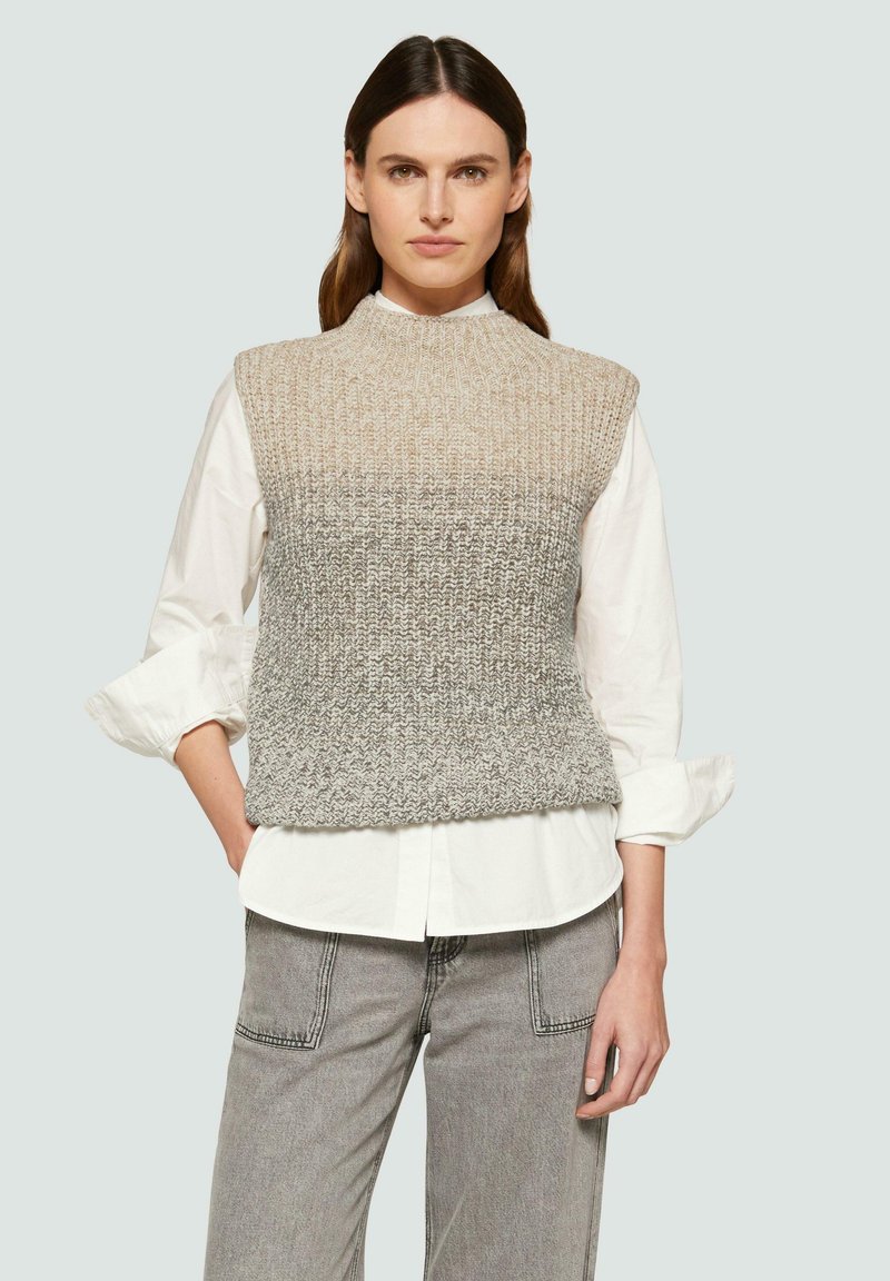 Gehaakte mouwloze trui in een beige naar grijs ombré vervaging, met een hoge hals en textuurafwerking, gedragen over een witte blouse met kraag.