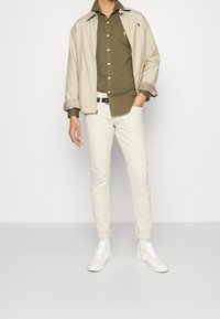 Giacca leggera beige sopra una camicia verde oliva, pantaloni cream slim e sneakers bianche. Dettagli notevoli includono le maniche arrotolate e una cintura nera.