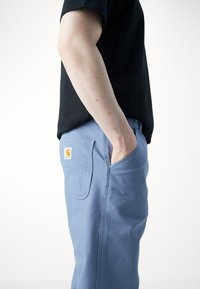 Lichtblauwe broek met een zijzak en een grote achterzak, voorzien van een geel merklogo. Gecombineerd met een zwart T-shirt.