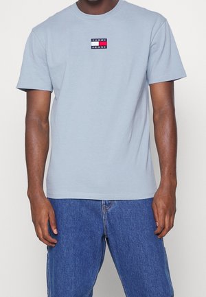 Mężczyzna ubrany w jasnoniebieski t-shirt Tommy Jeans i niebieskie dżinsy, stoi z rozluźnionymi ramionami wzdłuż boków na jednolitym tle.