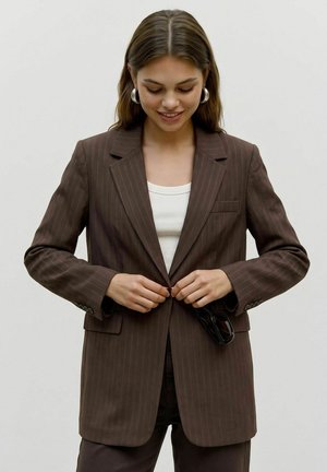 Jeune femme portant une veste de costume marron à fines rayures en train de la boutonner tout en tenant des lunettes de soleil noires, vêtue d'un haut blanc et de boucles d'oreilles créoles, souriant vers le bas.