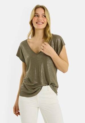Femme souriante aux longs cheveux blonds portant un haut vert olive brillant à col en V et manches courtes, et un pantalon blanc, tenant ses cheveux près de l'épaule.