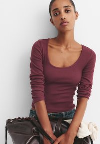 Maglietta a maniche lunghe a coste color bordeaux con scollatura a barca, abbinata a jeans blu e una borsa in pelle marrone scuro con dettagli rossi.