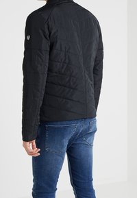 Czarna pikowana kurtka z wysokim kołnierzem, z logo na rękawie i dopasowanym kroju. Noszona z niebieskimi dżinsami.
