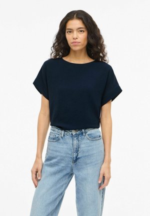 Žena s kučeravými vlasmi, ktorá má na sebe krátky námornícky modrý top zastrčený do svetlomodrých džínsov s vysokým pásom, na bielom pozadí.