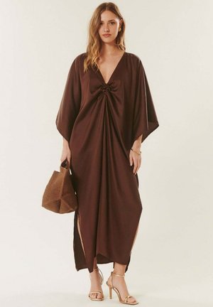 Femme portant une longue robe marron ample avec un col en V profond, des fentes sur les côtés, tenant un sac à main marron texturé et portant des sandales à talons hauts dorées.